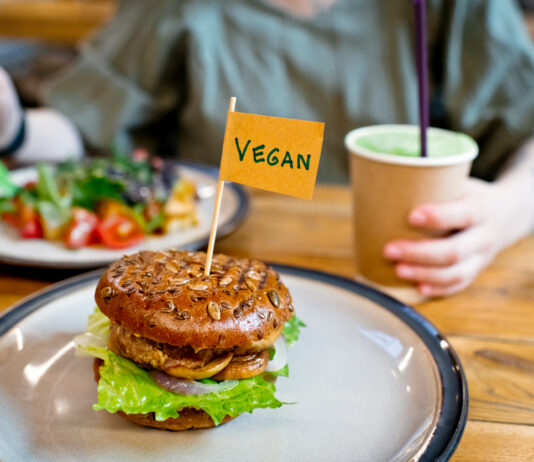 Eine Frau isst einen Salat und vor ihr auf einem weiteren Teller befindet sich ein Veggie Burger. Sie ernährt sich offensichtlich vegan und trinkt aus einem Coffee-to-go-Becher.