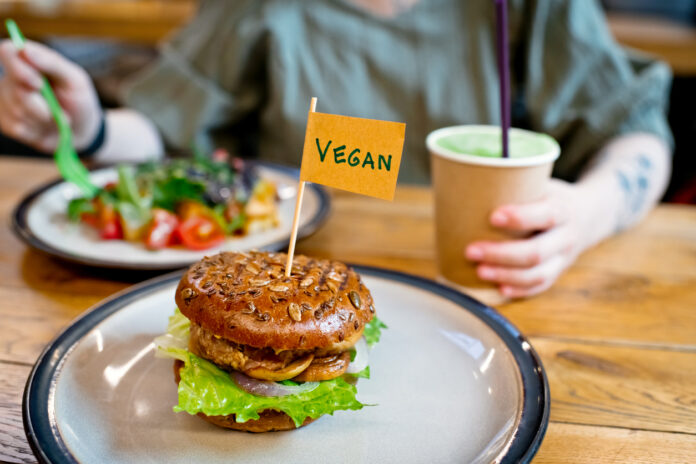 Eine Frau isst einen Salat und vor ihr auf einem weiteren Teller befindet sich ein Veggie Burger. Sie ernährt sich offensichtlich vegan und trinkt aus einem Coffee-to-go-Becher.