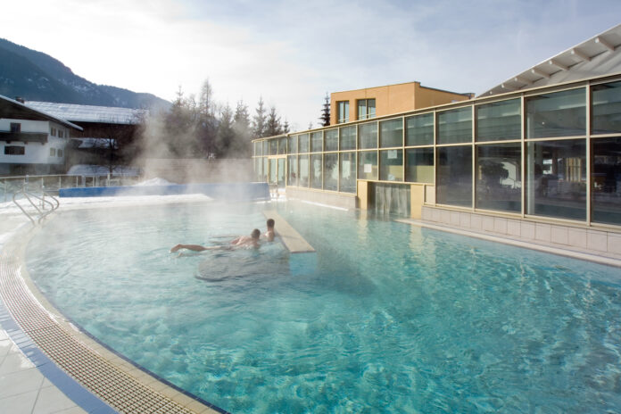 Zwei Männer in der Therme. Ein großes Außenschwimmbecken ist zu sehen, aus dem Dampf aufsteigt. Die Therme hat einen Innenbereich und einen Außenbereich, in dem sich die Gäste erholen und schwimmen können.