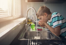 Ein kleiner Junge trinkt Wasser aus dem Wasserhahn in der Küche. Er schöpft das Wasser direkt mit den Händen aus dem Hahn und führt diese zum Mund.