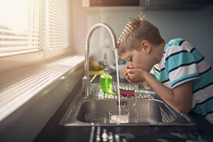 Ein kleiner Junge trinkt Wasser aus dem Wasserhahn in der Küche. Er schöpft das Wasser direkt mit den Händen aus dem Hahn und führt diese zum Mund.