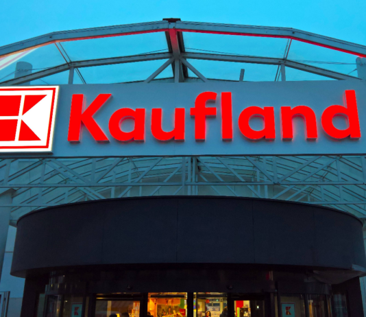 Der große Eingang einer Filiale von Kaufland. Der Standort erscheint gigantisch. Über den Eingangstüren prangt in roter Schrift der Kaufland-Schriftzug. Links daneben leuchtet das berühmte Logo der Supermarkt-Kette.