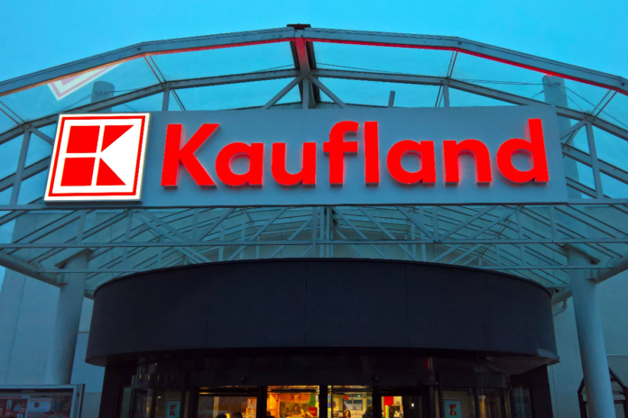 Der große Eingang einer Filiale von Kaufland. Der Standort erscheint gigantisch. Über den Eingangstüren prangt in roter Schrift der Kaufland-Schriftzug. Links daneben leuchtet das berühmte Logo der Supermarkt-Kette.