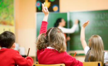 Klassenzimmer mit Kindern. Die Jungen und Mädchen sitzen an den Tischen im Unterricht und hören der Lehrerin zu. Diese schreibt gerade etwas an die Tafel mit Kreide.