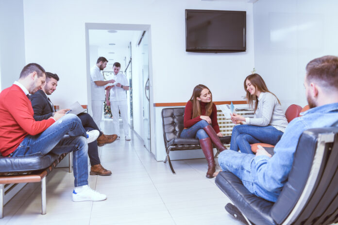Mehrere Patienten, Frauen und Männer, sitzen auf Bänken und Stühlen in einem Wartezimmer. Ärzte besprechen sich bei offener Tür einen Raum weiter, während die wartenden Patienten lesen, sich unterhalten oder einfach warten.