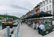 Touristen schlendern an einem Flussufer entlang. Im Hafen liegt ein schönes Schiff. Cafébesucher sitzen in einem Café und essen und trinken unter schwarzen Sonnenschirmen.