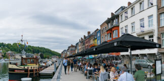 Touristen schlendern an einem Flussufer entlang. Im Hafen liegt ein schönes Schiff. Cafébesucher sitzen in einem Café und essen und trinken unter schwarzen Sonnenschirmen.