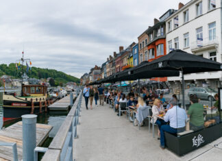 Touristen schlendern an einem Flussufer entlang. Im Hafen liegt ein schönes Schiff. Cafébesucher sitzen in einem Café und essen und trinken unter schwarzen Sonnenschirmen.