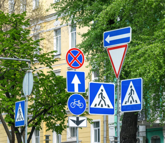 Vor einer Hausfassade sind viele verschiedene Verkehrszeichen zu sehen, die alle auf einmal gelten sollen. Für Verkehrsteilnehmer ist dies besonders verwirrend. Dies soll offensichtlich den Schilderwahn darstellen, der mancherorts herrscht.