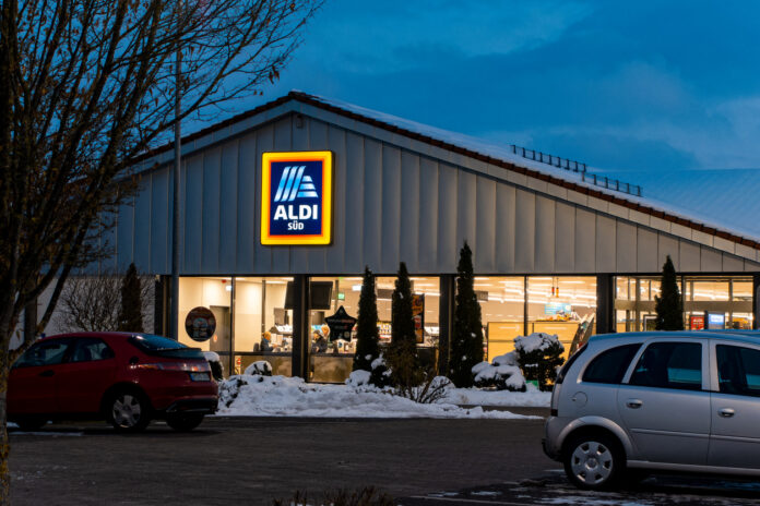 Eine Filiale von Aldi Süd steht in der Abenddämmerung. Es ist schon dunkel, daher ist bald Ladenschluss. Im Vordergrund auf dem Parkplatz stehen zwei Autos, und es liegt ein bisschen Schnee.