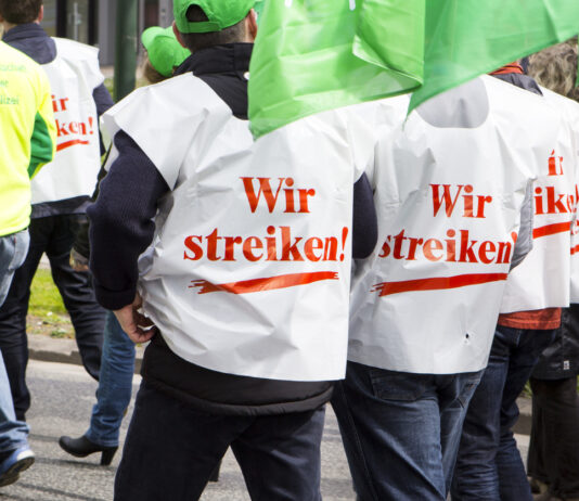 Mehrere Demonstranten laufen in Shirts mit der Aufschrift "wir streiken" durch die Stadt im Zusammenhang mit einer Demonstration. Man sieht die Menschen nur von hinten, aber nicht ihre Gesichter.