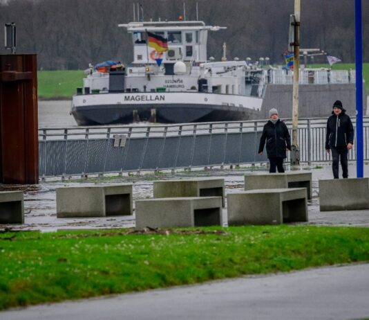 Ein Schiff mit dem Namen „Malgellan“ fährt auf dem Karlsruher Rhein entlang. Am Ufer entlang spazieren zwei Personen mit Mützen. Es ist offenbar kalt und regnet.