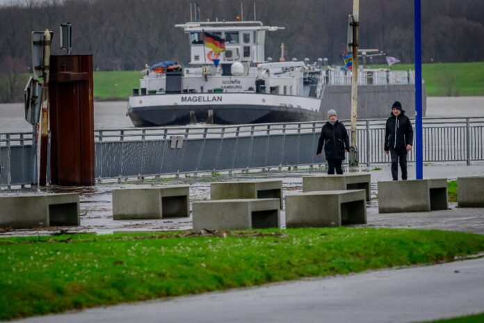 Ein Schiff mit dem Namen „Malgellan“ fährt auf dem Karlsruher Rhein entlang. Am Ufer entlang spazieren zwei Personen mit Mützen. Es ist offenbar kalt und regnet.