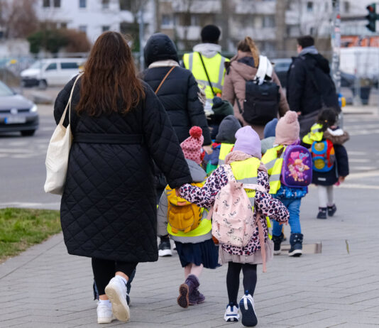 Einige Kindergartenkinder laufen mit ihren Erziehern an einer Straße entlang. Die Kinder tragen alle eine gelbe Warnweste. Es werden nun heftige Maßnahmen erwartet.