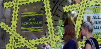 Geschlossenes Geschäft in der Innenstadt. Zwei Kundinnen stehen vor einem abgeriegelten Laden. Auf einem dunklen Schild steht. Wir schließen. Das Schaufenster ist leer und nicht mehr befüllt.