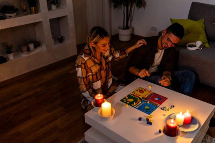 Ein Paar sitzt vor einem Tisch, auf dem brennende Kerzen stehen und spielen ein Gesellschaftsspiel. Der Strom scheint nicht mehr zu funktionieren, wie bei einem Stromausfall.