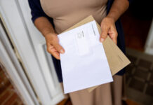 Eine Frau steht in ihrer geöffneten Haustür und hält die Post in der Hand. Sie liest gerade einen Brief, den sie aus einem braunen Umschlag genommen hat.