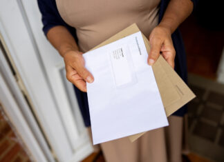 Eine Frau steht in ihrer geöffneten Haustür und hält die Post in der Hand. Sie liest gerade einen Brief, den sie aus einem braunen Umschlag genommen hat.