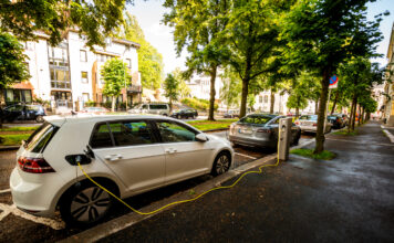 Ein E-Auto lädt an einer Ladesäule neben der Straße. Aus dem Tankdeckel ragt ein gelbes Kabel. Dahinter sind Bäume zu sehen.