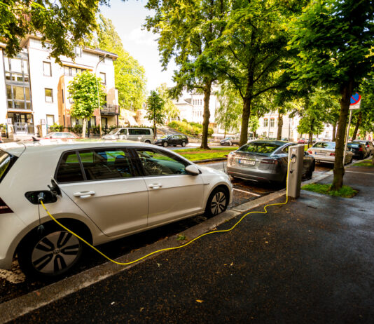 Ein E-Auto lädt an einer Ladesäule neben der Straße. Aus dem Tankdeckel ragt ein gelbes Kabel. Dahinter sind Bäume zu sehen.