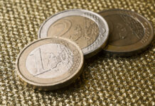 Zwei Ein-Euro-Stücke und ein Zwei-Euro-Stück liegen auf einer goldenen und strukturierten Oberfläche. Das Bargeld ist Gold und Silber und aufeinander gestapelt.