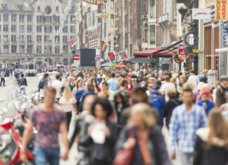 Zahlreiche Menschen drängen sich in einer Einkaufsstraße mitten in der Innenstadt. Das Foto ist verwaschen, jedoch erkennt man viele junge und alte Bürger sowie dutzende Geschäfte, Fassenden, Cafés und den gesamten Trubel der Straße in der Innenstadt.