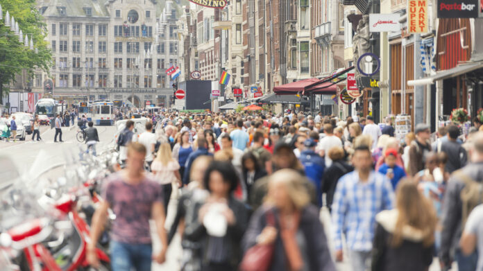 Zahlreiche Menschen drängen sich in einer Einkaufsstraße mitten in der Innenstadt. Das Foto ist verwaschen, jedoch erkennt man viele junge und alte Bürger sowie dutzende Geschäfte, Fassenden, Cafés und den gesamten Trubel der Straße in der Innenstadt.