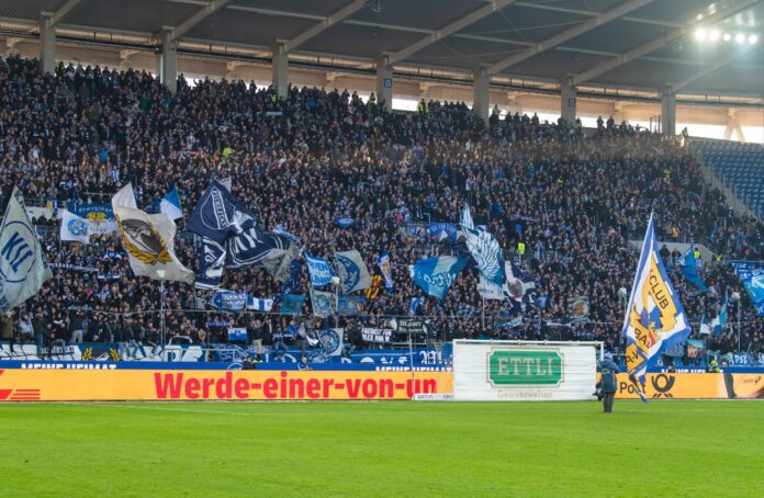 Die Fans des Karlsruher SC jubeln auf der Tribüne vor ihnen der Rasen des Fußballstadions