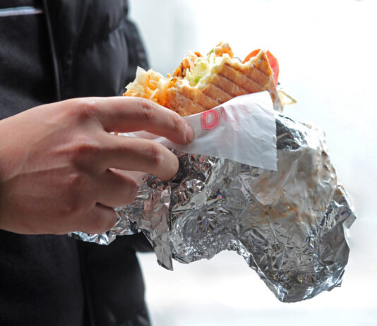 Ein Mann hält einen Döner in der Hand. Der Döner ist schon zur Hälfte aufgegessen und das Silberpapier ist halb abgewickelt.