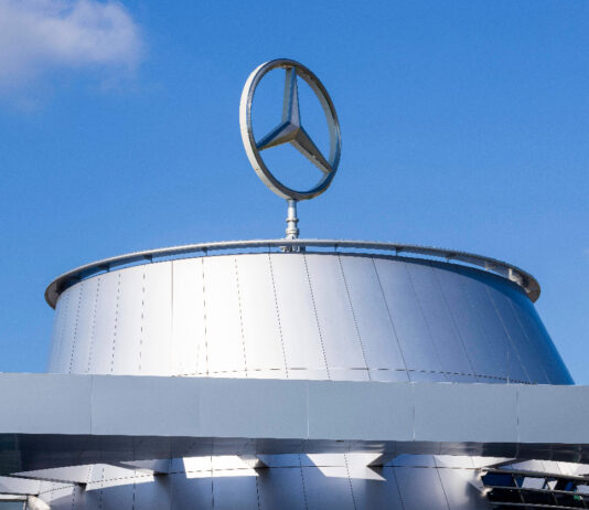 Das Gebäude einer Mercedes-Niederlassung mit dem Mercedes-Stern auf dem Dach. Das Firmen-Logo prangt am blauen Himmel bei sonnigem Wetter. Die Niederlassung könnte ein Werk oder eine Zentrale sein.