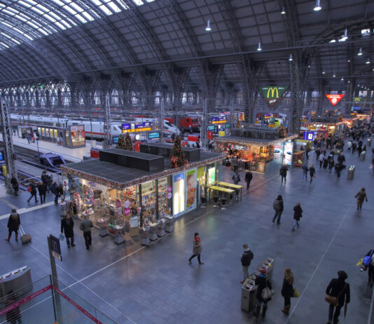 Viele Menschen befinden sich in einer Bahnhofshalle. Im Hintergrund sind verschiedene Geschäfte und Restaurants. Der Bahnhof hat eine gläserne Überdachung.