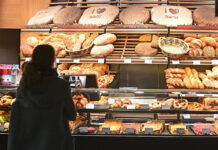 Eine Kundin steht in einer Bäckerei vor einer Auslage von verschiedenen Backwaren wie Brot, Brötchen und Süßgebäck. Viele Bäckereien führen jetzt eine Zusatzgebühr ein.