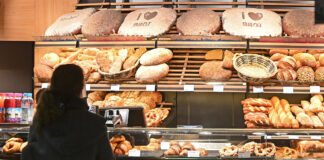 Eine Kundin steht in einer Bäckerei vor einer Auslage von verschiedenen Backwaren wie Brot, Brötchen und Süßgebäck. Viele Bäckereien führen jetzt eine Zusatzgebühr ein.