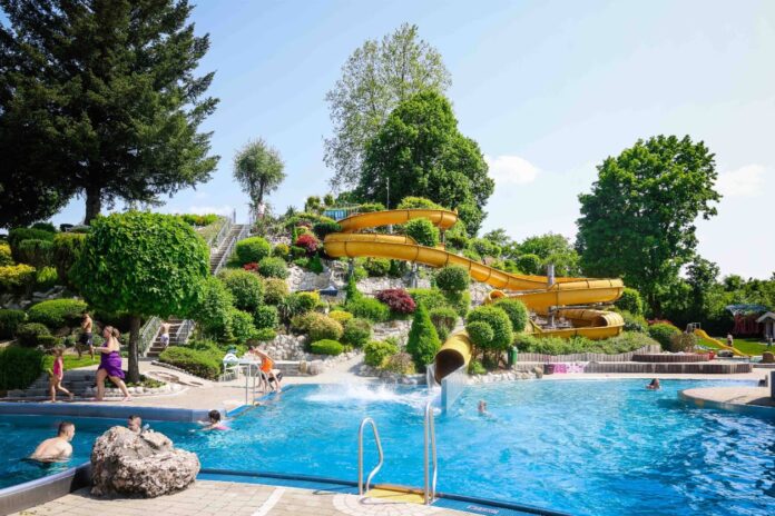 Ein Spaßbad oder ein Freibad im Sommer mit vielen Besuchern, darunter Kinder und Erwachsene. Es gibt ein Schwimmbecken und einen Nichtschwimmerbereich. Im Hintergrund befindet sich eine Wasserrutsche.