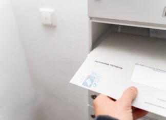 Eine Hand nimmt einen offiziell wirkenden Brief samt Briefumschlag aus dem Briefkasten. Ein Logo auf dem Umschlag verrät, dass es sich wohl um amtliche Post oder etwas von einer staatlichen Behörde handeln muss.