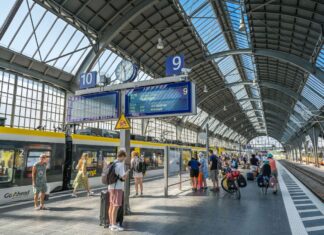 Ein überdachter Bahnsteig eines offensichtlich großen Bahnhofs, sehr wahrscheinlich eines Hauptbahnhofs. Viele Kinder und andere Reisende warten auf ihre Züge.