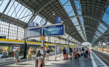 Ein überdachter Bahnsteig eines offensichtlich großen Bahnhofs, sehr wahrscheinlich eines Hauptbahnhofs. Viele Kinder und andere Reisende warten auf ihre Züge.