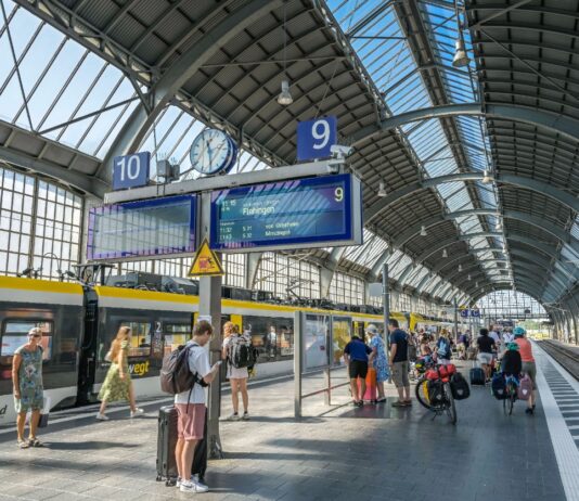 Ein überdachter Bahnsteig eines offensichtlich großen Bahnhofs, sehr wahrscheinlich eines Hauptbahnhofs. Viele Kinder und andere Reisende warten auf ihre Züge.