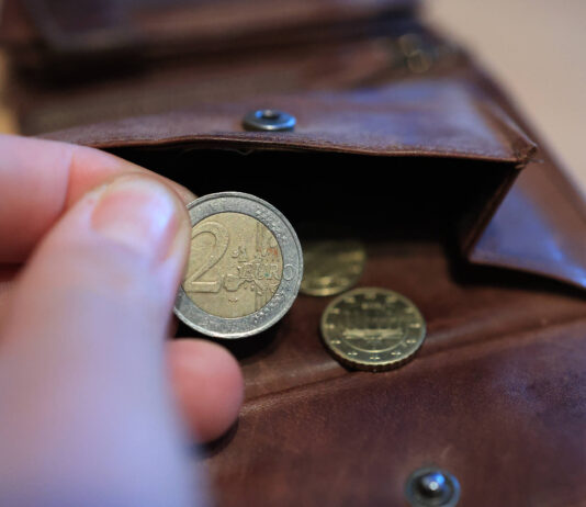 Eine Hand hält eine 2-Euro-Münze, die eine Person aus dem Portemonnaie genommen hat. Weitere Münzen befinden sich im Geldbeutel.