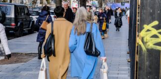 Zwei Frauen mit Einkaufstüten spazieren durch eine Innenstadt. Im Hintergrund sieht man viele weitere Menschen, die einen Einkaufsbummel durch die Fußgängerzone machen.