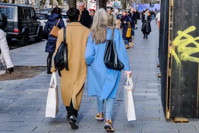 Zwei Frauen mit Einkaufstüten spazieren durch eine Innenstadt. Im Hintergrund sieht man viele weitere Menschen, die einen Einkaufsbummel durch die Fußgängerzone machen.