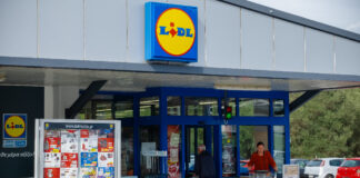 Der Eingang einer Lidl-Filiale mit geöffneten Türen und mit einem Schaukasten mit den aktuellen Angeboten der Woche und dem Parkplatz im Hintergrund. Lidl und Kaufland ändern nun etwas Traditionelles an der Kasse.