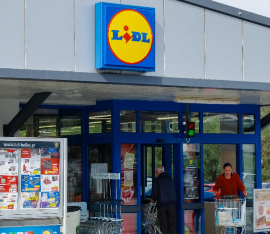 Der Eingang einer Lidl-Filiale mit geöffneten Türen und mit einem Schaukasten mit den aktuellen Angeboten der Woche und dem Parkplatz im Hintergrund. Lidl und Kaufland ändern nun etwas Traditionelles an der Kasse.