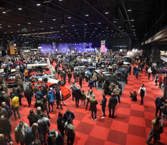 In einer Ausstellungshalle für Oldtimer verschiedener Automarken tummeln sich viele Menschen, um sich die Autos anzusehen. In der Halle befinden sich, gut ausgeleuchtet, viele verschiedene Modelle.