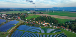 Luftaufnahme eines sehr großen Solarfeldes auf einem Acker. Am Rand des Feldes lässt sich ein Dorf erkennen. Es handelt sich dabei offenbar um eine Landschaft auf deutschem Boden. Im Hintergrund lassen sich zudem Windräder erkennen.