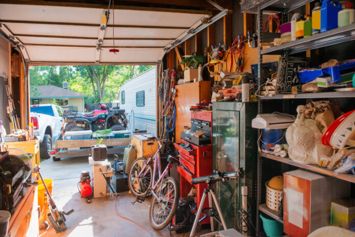 Der Blick in eine Garage, die über und über vollgestellt ist. Sämtliche Regale sind voll gepackt mit den unterschiedlichsten Dingen. Man kann ein Fernrohr, aber auch ein Fahrrad erkennen. Der Blick nach draußen zeigt, dass dort ein Haus sowie ein Campingwagen steht.