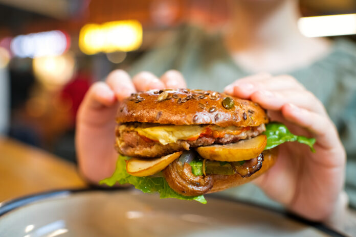 Eine Frau isst in einem Restaurant einen Veggie-Burger. Dieser Hamburger ist sehr vegetarisch, aber nicht vegan. Das bedeutet: Es ist kein tierisches Fleisch enthalten.