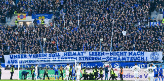 Das Stadion des Karlsruher SC. Die Spieler stehen auf dem Platz und lassen sich von ihren Tausenden Fans in Weiß und Blau feiern – gut sichtbar ist ein Banner für den KSC am Spielfeldrand angebracht.