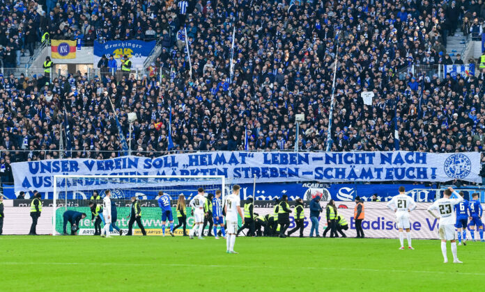 Das Stadion des Karlsruher SC. Die Spieler stehen auf dem Platz und lassen sich von ihren Tausenden Fans in Weiß und Blau feiern – gut sichtbar ist ein Banner für den KSC am Spielfeldrand angebracht.