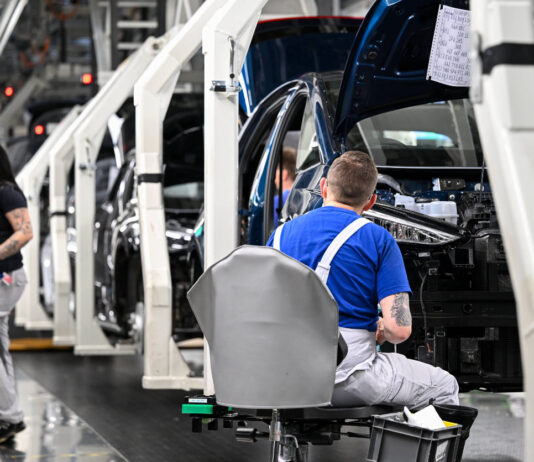 Eine Produktionsstraße für Autos mit Mitarbeitern. In der Halle der Fabrik setzen Arbeiter Teile der Autos zusammen. Ein Arbeiter kontrolliert die Fahrzeugfront. Am linken Bildrand stehen noch zwei Angestellte in Arbeitskluft. Ein deutscher Autobauer will nun massiv Stellen abbauen.